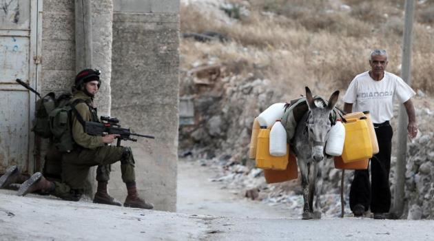 Palestinian man leads a donkey carrying water jerrycans as Israeli soldiers take position during an army search operation in the West Bank village of Awarta/AFP-Getty